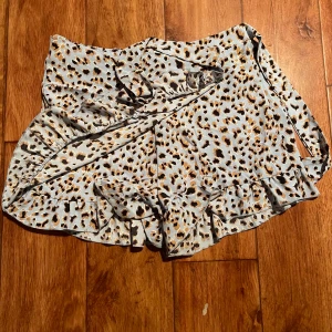 Shorts med volang - Säljer ett par shorts med omlott på framsidan med leopardmönster i beige, svart och brunt. Shortsen har volangdetalj längs nederkanten och knyts i sidan för en snygg passform. Perfekt för dig som vill sticka ut med ett trendigt djurmönster.