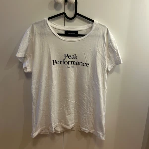 Vit Peak Performance t-shirt i bomull - Vit t-shirt från Peak Performance med svart tryck på bröstet. Klassisk rund hals och korta ärmar, tillverkad i mjuk bomull. Perfekt för dig som gillar enkel och stilren design med en sportig touch.