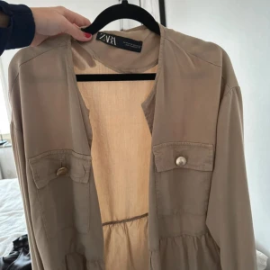 Beige klänning med fickor från Zara - Snygg beige klänning från Zara med långa ärmar och två stora fickor framtill med dekorativa knappar. Klänningen har en avslappnad passform och markerad midja med rynkade detaljer. Perfekt för dig som gillar stilrena och enkla plagg.
