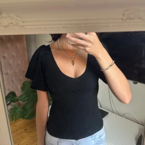 Svart ribbad topp med knyt i ryggen - Säljer en svart ribbad topp med v-ringning och korta, vida ärmar. Toppen har en snygg öppen rygg med knytband upptill som ger en extra detalj. Figurnära passform och stretchigt material.