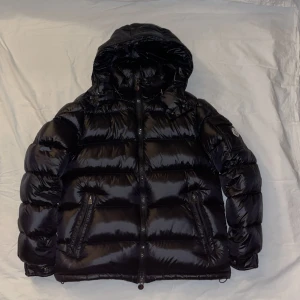 Moncler Maya - Snygg svart dunjacka från Moncler med huva och dragkedja. Jackan har ett glansigt yttre och Moncler-logga på ärmen. Perfekt för kyliga dagar.