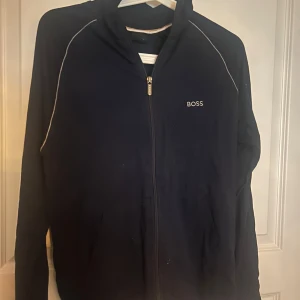 blå ziptröja från BOSS - Säljer en blå långärmad ziptröja från BOSS med vit piping längs axlarna och diskret logga på bröstet. Tröjan har hög krage och dragkedja framtill, perfekt för lager-på-lager. Materialet känns mjukt och bekvämt.