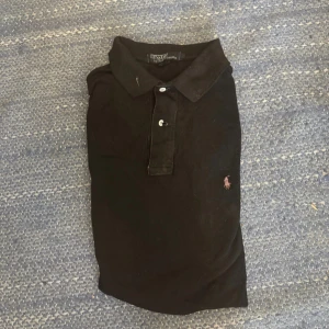 Svart pikétröja från Polo Ralph Lauren - Svart pikétröja från Polo Ralph Lauren med klassisk krage och två vita knappar. Tröjan har det ikoniska broderade Polo-logot i rosa på bröstet. Tillverkad i mjuk bomull som är skön mot huden och har korta ärmar. Perfekt för en clean och stilren look.