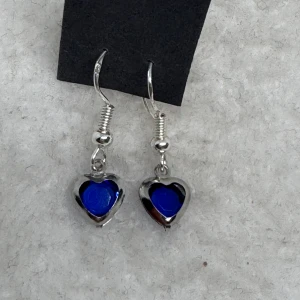 Blå hjärtformade örhängen i silver - Supersöta örhängen med hjärtformad blå sten infattad i silverfärgad metall. Hängande modell med krok, perfekt för att ge din look en färgklick och lite extra kärlek. Krokarna är i sterling silver. Nya