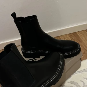 Svarta chelsea boots från ONLY - Snygga svarta chelsea boots från ONLY med grov sula och vit kontrastsöm runt sulan. Skorna är i skinnimitation och har elastiska paneler på sidorna för enkel påtagning. Perfekta för dig som vill ha en stilren och trendig look.
