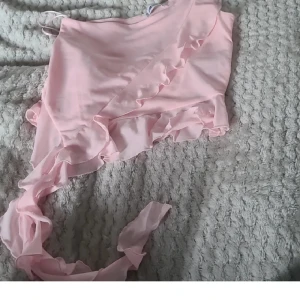 Ljusrosa omlottopp med volang - Supersöt ljusrosa omlottopp med tunna axelband och volangdetaljer längs framsidan och nederkanten. Toppen har en lång knytband som ger en snygg och feminin look. Perfekt för dig som gillar romantiska vibes och pastellfärger.