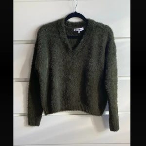 Mörkgrön fluffig stickad tröja - Mysig mörkgrön stickad tröja med fluffig textur och v-ringad hals. Perfekt för kyliga dagar och enkel att styla med jeans eller kjol. Långärmad modell med avslappnad passform.