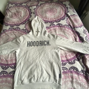 Vit HOODRICH hoodie med tryck - Säljer en vit hoodie från HOODRICH med svart tryck över bröstet. Klassisk känguruficka framtill, huva med snörhål och långa ärmar. Perfekt för dig som gillar streetwear och vill ha en enkel men snygg look.