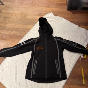 Svart vindjacka Rebel med huva M - Svart vindjacka från Rebel med grå detaljer och reflexränder. Jackan har huva med dragsko, två dragkedjeförsedda fickor och broderad orange logga på bröstet. Tillverkad i polyester och spandex för smidig passform. Perfekt för blåsiga dagar. Bra skick ovanpå inte så bra inuti. De limmade banden på sömmarna har släppt. Se bild
