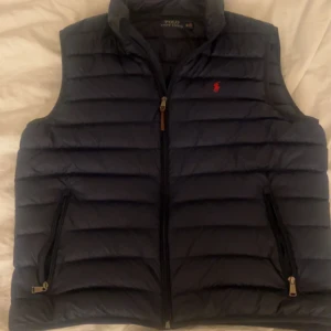 Mörkblå dunväst från Polo Ralph Lauren - Snygg mörkblå dunväst från Polo Ralph Lauren med horisontella quiltade sömmar och klassisk röd logga på bröstet. Västen har dragkedja framtill och två fickor med dragkedja. Perfekt för lager-på-lager-stil under kyliga dagar.