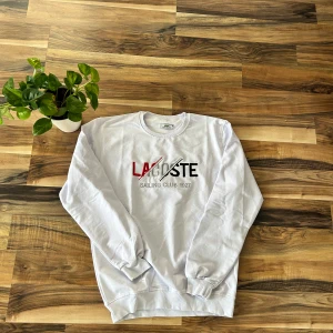 Vit sweatshirt från Lacoste S - Vit sweatshirt från Lacoste med broderad text i rött, svart och silver på bröstet där det står 'Lacoste Sailing Club 1927'. Tröjan har rund halsringning, långa ärmar och ribbade muddar. Perfekt för en clean och sportig stil.