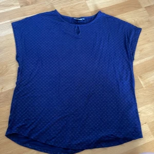 Mörkblå blus från Fransa - Säljer en mörkblå blus från Fransa med diskret mönster och en liten droppformad öppning vid halsen. Blusen har korta ärmar och är tillverkad i ett mjukt, lätt material som känns skönt mot huden. Perfekt för dig som gillar en enkel men snygg look. Färgen är mer mörkblå i verkligheten. 