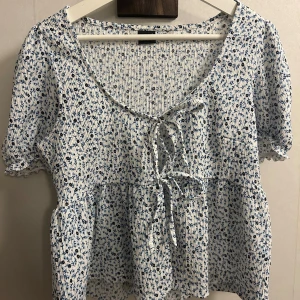 Blommig blus med knyt från Gina Tricot - Supersöt blommig blus från Gina Tricot i vitt och blått. Modellen har kort puffärm, v-ringning och knyt framtill. Tyget är ribbat och lätt, perfekt för vår och sommar. Blusen har volang nedtill och småblommigt mönster över hela plagget.