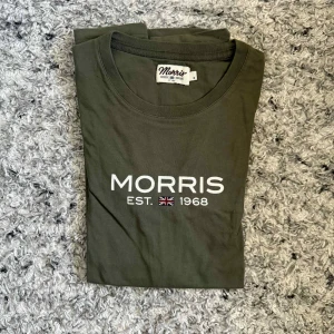 Olivgrön t-shirt från Morris - Snygg olivgrön t-shirt från Morris med vit logga och brittisk flagga tryckt på bröstet. Klassisk rund hals och korta ärmar. Tillverkad i mjuk bomull som känns skön mot huden. Perfekt för en clean och stilren look.