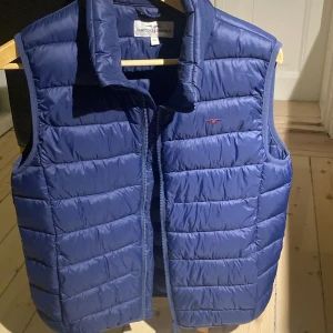 Hampton Republic väst - Snygg blå dunväst från Hampton Republic med quiltad design och hög krage. Västen har dragkedja framtill och en liten broderad logga på bröstet. Perfekt att slänga över en hoodie för en cool och sportig look.