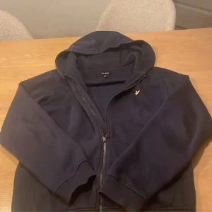 Mörkblå hoodie från Lyle & Scott - Säljer en mörkblå hoodie från Lyle & Scott med dragkedja och huva. Tröjan har det klassiska gula fågel-loggan broderad på bröstet och är tillverkad i mjuk bomull. Perfekt för dig som gillar stilrena och bekväma plagg.