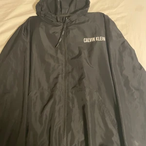 Svart vindjacka från Calvin Klein - Svart vindjacka från Calvin Klein med huva och dragkedja framtill. Jackan har logga tryckt på bröstet och två fickor på sidorna. Tillverkad i lätt polyester, perfekt för blåsiga dagar. Enkel och stilren design som funkar till det mesta.