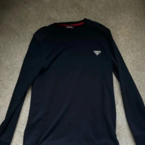 Mörkblå långärmad tröja från Prada - Snygg mörkblå långärmad tröja från Prada med diskret logga på bröstet och coola detaljer på axlarna. Tröjan har rund halsringning med röd insida och är i ett mjukt material som känns skönt mot huden. Perfekt för dig som gillar stilrena designer.