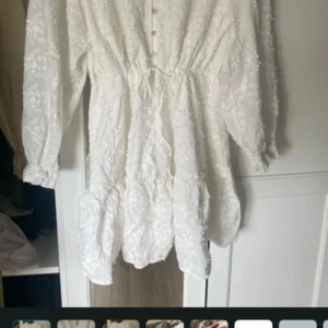 Vit Suri dress med spets och knyt - Superfin vit kortklänning från Suri med broderade blommor, spetsdetaljer och knyt i midjan. Klänningen har långa ärmar, knappar framtill och volang nertill. Perfekt för sommarens alla tillfällen när du vill känna dig extra söt. Använd endast en gång 