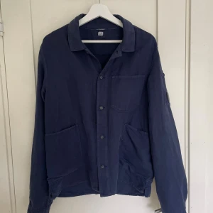 Marinblå overshirt från C.P. Company - Säljer en stilren blå overshirt från C.P. Company med klassisk krage, stora fickor framtill och ikonisk linsdetalj på ärmen. Jackan har knäppning med knappar. Perfekt för lager-på-lager och snygg till jeans. Använd en gång men säljs pågrund av att den är för stor.