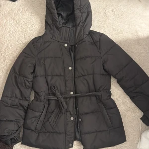 Svart dunjacka från Nelly - Svart pufferjacka med huva och knytbälte i midjan. Jackan är köpt från Nelly och är i storlek xs