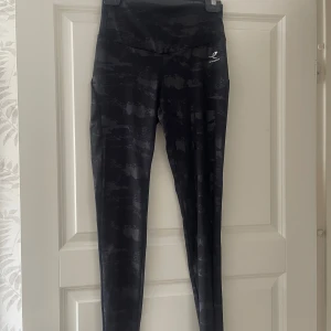 Svarta camo leggings  - Snygga svarta leggings med mörkgrått camo-mönster. Hög midja och tight passform, perfekta för träning eller chill. Tillverkade i stretchigt syntetmaterial som andas och sitter skönt hela dagen.