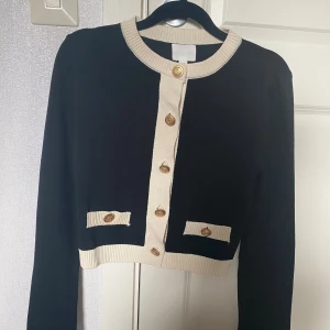 Svart och beige kofta - Stilren svart kofta med beigea detaljer och guldknappar framtill, med långa ärmar och dekorativa fickor. Perfekt för dig som gillar klassisk men trendig look.