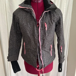 Vinterjacka  från Woxnerud - Grå vinterjacka med svart leopardmönster och rosa detaljer från Wox. Jackan har huva, flera fickor med rosa dragkedjor och svarta muddar med tumhål. Insidan är quiltad och har borttagbart snölås. Tyvärr ser de ljusrosa delarna lite dassiga ut, men jackan har mer kvar att ge. (Diskret namnad på insidan)