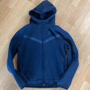 Svart Nike tech hoodie med dragkedja - Snygg Nike tech hoodie i storlek 158-170 cm. Tröjan har hel dragkedja framtill, huva och diskret Nike-logga på bröstet. Tillverkad i mjukt material, perfekt för chill eller träning. Klassisk design med långa ärmar och bekväm passform.