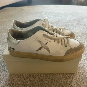 Vit sneaker från Axel Arigato, stl 40 - Snygga vita sneakers från Axel Arigato med broderad fågel på sidan och texten 'ARIGATO'. Skorna har beige sula, silverdetaljer vid hälen och är tillverkade i skinn. Klassisk rund tå och snörning framtill. Handgjorda i Portugal.