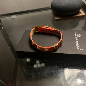 Liknade valentino armband  - Säljer ett orange armband från Denuasi i skinn med coola pyramidformade guldfärgade nitar. Armbandet har justerbart spänne och en snygg, trendig look som sticker ut. Perfekt accessoar för dig som vill ha något edgy och unikt.