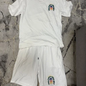 Vita shorts och t shirt från Casablanca med tryck - Säljer ett par vita shorts och t shirt  från Casablanca med färgglatt tryck och texten 'Avenida Casablanca' på ena benet. Shortsen har snörning i midjan och är gjorda i mjuk bomull, perfekta för sommardagar. Matcha med t-shirt för en clean och chill look.