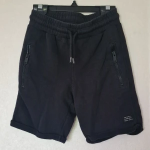 Svarta shorts från Kappahl - Svarta shorts från Kappahl med elastisk midja och snörning. Två fickor framtill med dragkedjor och en bakficka. Perfekta för chill dagar eller när du vill ha en avslappnad stil.