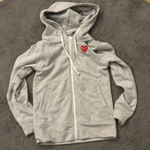  Comme des Garçons Play CDG - Grå hoodie från Comme des Garçons Play med dragkedja och huva. Ikoniskt rött hjärta med ögon broderat på bröstet. Mjuk bomullskvalitet, två fickor fram och vita snören i huvan. Perfekt för en chill och stilren look.