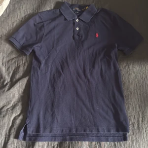 Mörkblå Polo Ralph Lauren t-shirt - Snygg mörkblå t-shirt från Polo Ralph Lauren med klassisk krage och tre vita knappar. Den har korta ärmar och röd broderad logga på bröstet. Tillverkad i mjuk bomull som känns skön mot huden. Passformen är slim och ger en stilren look.