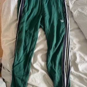 Gröna Adidas mjukisbyxor med ränder - Gröna mjukisbyxor från Adidas med klassiska vita och svarta ränder längs sidorna. Byxorna har resår i midjan och vid bensluten samt broderad Adidas-logga på låret. Tillverkade i mjukt material som är skönt att ha på sig.