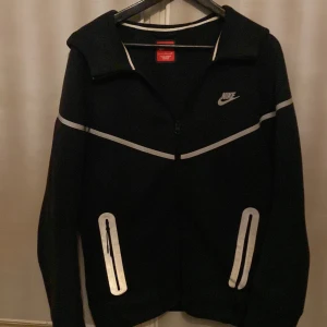 Svart Nike hoodie med reflexdetaljer - Svart hoodie från Nike med vita reflexdetaljer på bröstet och vid fickorna. Tröjan har dragkedja framtill, huva och långa ärmar. Perfekt för kvällspromenader eller träning när du vill synas. Snygg sportig look med klassisk Nike-logga på bröstet.