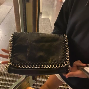 Tiamo väska - Snygg svart axelväska i skinn med grova silverfärgade kedjor längs kanterna. Väskan har lock och flera fack inuti, inklusive dragkedjefack. Perfekt storlek för det viktigaste och ger en cool vibe till outfiten.