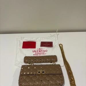 Valentino Garavani quiltad axelväska - Snygg beige axelväska från Valentino Garavani i quiltat skinn med guldiga nitar och kedja. Väskan har ett avtagbart axelband och klassiskt spänne framtill. Perfekt för dig som vill ha en statementväska med lyxig känsla. Hör av er vid frågor💕
