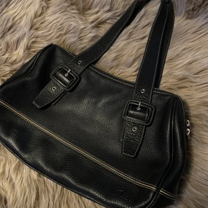 Svart handväska i skinn från Lacoste - Snygg svart handväska från Lacoste i äkta skinn med grov struktur. Väskan har dubbla axelband med spänndetaljer, kontrastsömmar och silverfärgade dragkedjor med Lacoste-logga. Klassisk krokodilprägling på framsidan och ljus insida med innerfack. Nyskick men kommmer inte till använding, därav säljs den. Liknande väskor från lacoste ligger på runt 2000-3000 nypris.