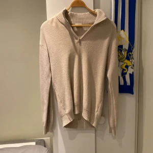 Beige stickad half zip-tröja Kappahl - Beige stickad tröja från Kappahl med ribbade muddar och krage. Tröjan har en snygg half zip-dragkedja framtill och är långärmad. Perfekt för lager-på-lager och chill höst- eller vinterstil.
