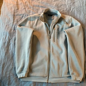 Ljusgrå ziptröja från Sail Racing - Ljusgrå långärmad ziptröja från Sail Racing med hög krage och dragkedja framtill. Tröjan har en bröstficka med dragkedja och broderad logga på bröstet. Tillverkad i mjuk bomullsmix, perfekt för lager-på-lager under kyliga dagar.