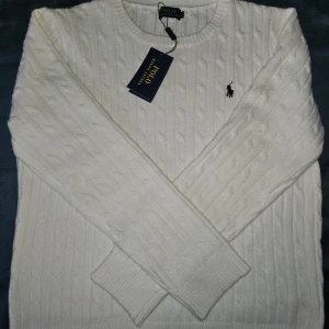 Vit kabelstickad tröja Polo Ralph Lauren - Vit kabelstickad tröja från Polo Ralph Lauren med klassisk rund halsringning och broderad logga på bröstet. Tröjan har långa ärmar och ribbade muddar, perfekt för dig som gillar stilren och tidlös design.