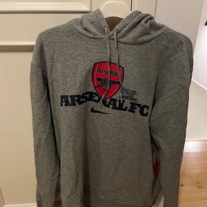 Grå Arsenal Nike hoodie - Grå hoodie från Nike med Arsenal FC-logga och tryck framtill. Tröjan har huva med röd insida och snörning, samt långa ärmar. Perfekt för dig som älskar fotboll och vill ha en sportig stil.