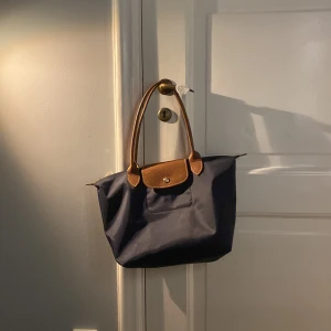 Mörkblå Longchamp Le Pliage axelväska - Snygg mörkblå Le Pliage axelväska från Longchamp med bruna skinnhandtag och detaljer🫶🏻🤭Väskan så fina har guldiga metalldetaljer och dragkedja🙂Du får plats med allt (dator, mappar m.m.) perfekt för dig som går i skolan och måste ta med dig allt till olika lektioner! Har små defekter i kanterna men verkligen inget man tänker på. I storlek medium. SKRIV OM FRÅGOR💞