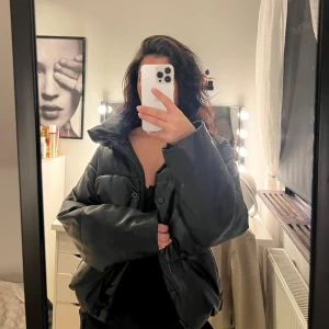 Svart pufferjacka - Svart pufferjacka med oversized passform och hög krage. Jackan har breda ärmar och stängs med både dragkedja och knappar. Materialet är glansig skinnimitation. Perfekt till höst och vinter. 