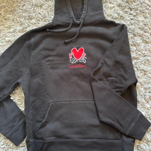 Svart hoodie Axel Arigato x Keith Haring - Svart hoodie från Axel Arigato med Keith Haring-broderi på bröstet. Tröjan har en röd hjärtgrafik, dragsko i huvan och känguruficka framtill. Mjuk bomullskvalitet och ribbade muddar. Märkespatch nertill på ryggen.