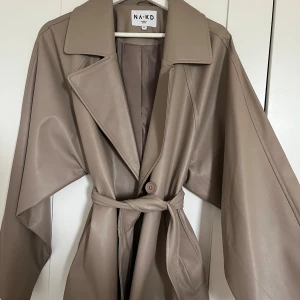 Beige trenchcoat i skinnimitation NA-KD - Snygg beige trenchcoat från NA-KD i skinnimitation med bred krage, knytskärp i midjan och långa ärmar. Jackan har en stilren look med knappar framtill och släta fickor. Kappan går ner till höfterna och passar som en oversized jacka. Materialet är som en regnjacka vilket är bra inför hösten. 