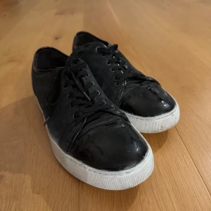 Svarta sneakers från Lanvin i mocka - Snygga svarta sneakers från Lanvin med ovandel i mocka och detaljer i blankt skinn. Skorna har vita, platta sulor och klassisk snörning framtill. Perfekta för dig som gillar stilrena och exklusiva sneakers med en twist. De är använda och på ena skon finns ett litet vitt märke. Detta bör synas på bilden. Nypris 5500 kr. 