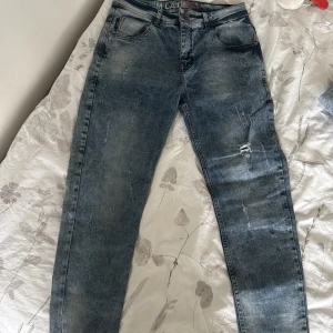 Blå jeansbyxor med slitningar - Säljer ett par blå jeansbyxor från Denim Lation med snygga slitningar och tvättad look. Klassisk femficksmodell med raka ben och normal passform. Jeansen har detaljerade bakfickor och är tillverkade i robust denim för en cool vardagsstil.